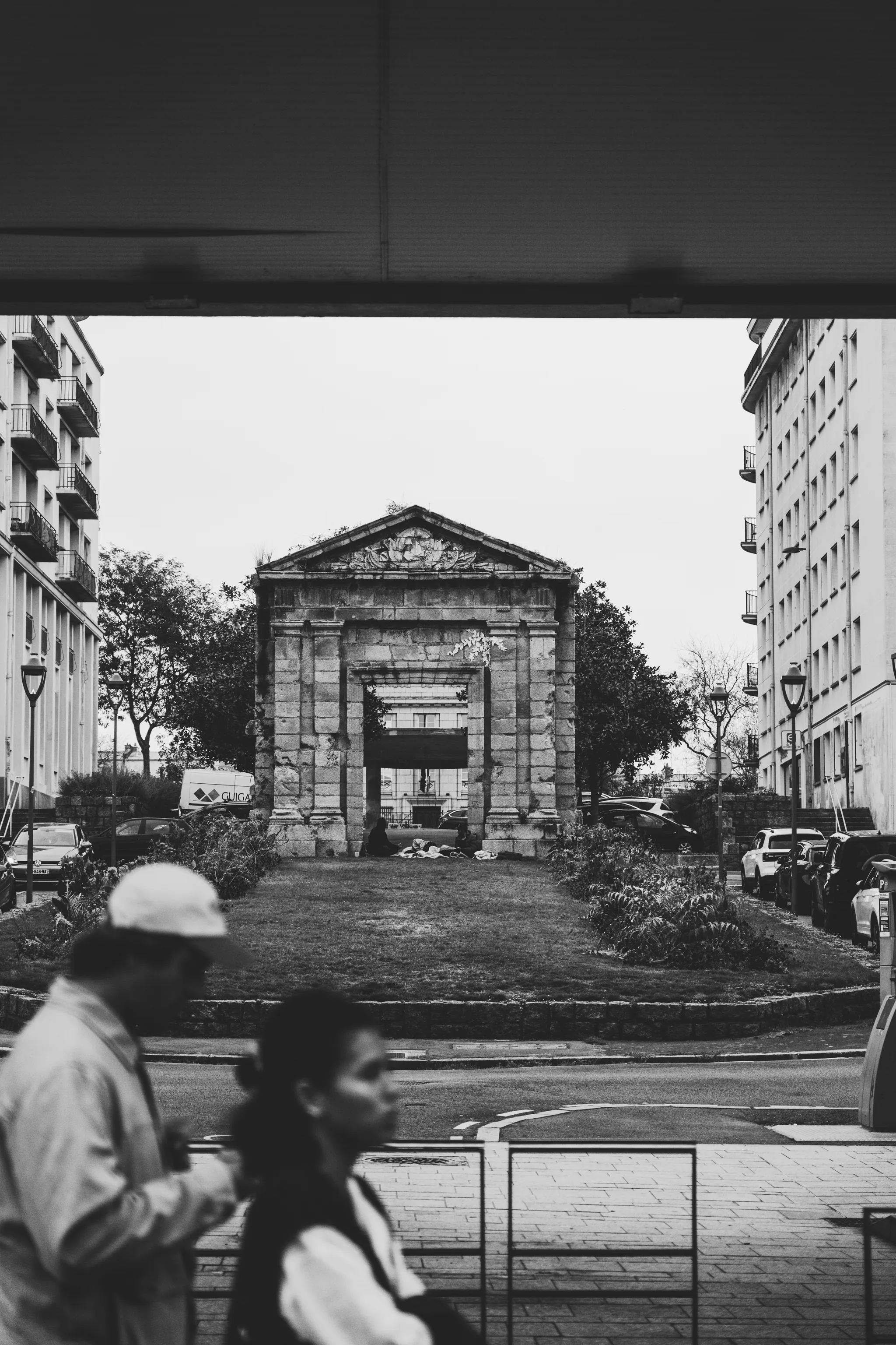 Stone gateway with a pediment standing alone in a small park between apartment blocks, passers-by in front, black and white
