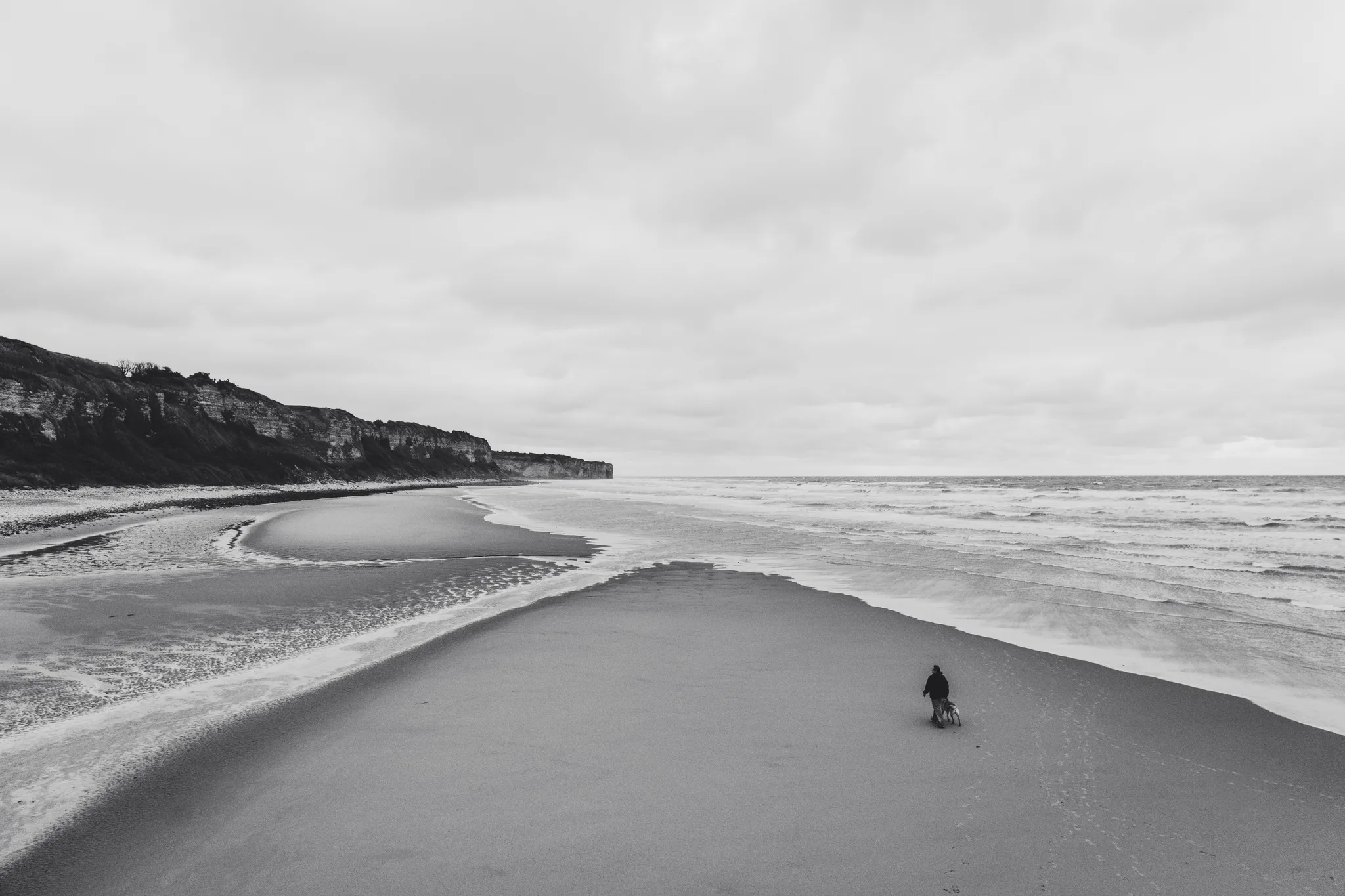 A lone figure on a long sand spit with cliffs behind, black and white