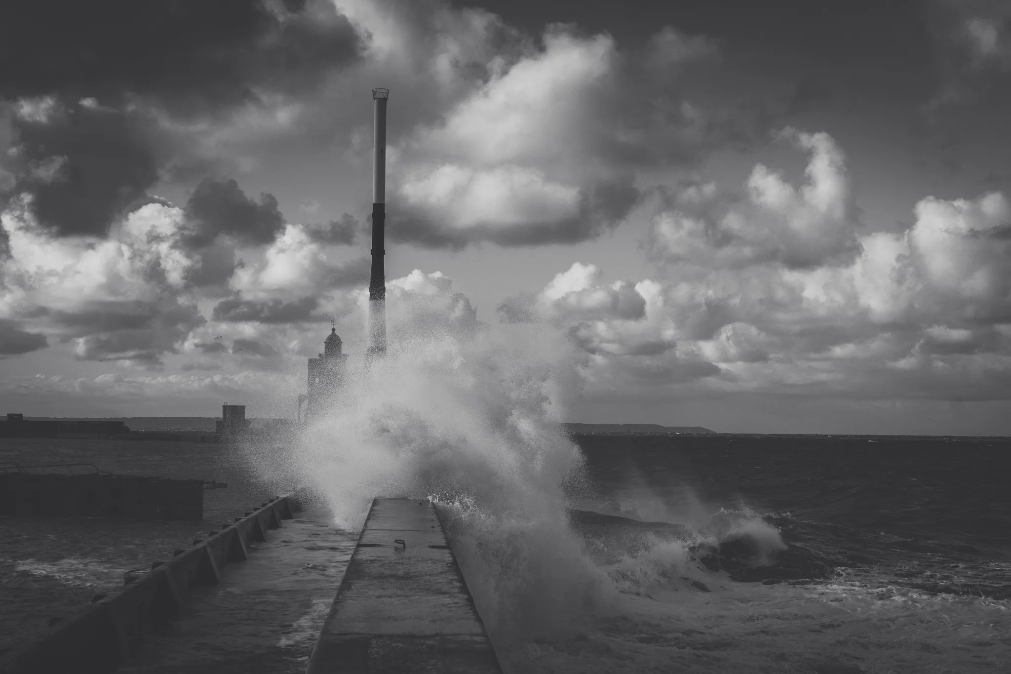 Wave exploding over a jetty in front of a domed lighthouse, black and white