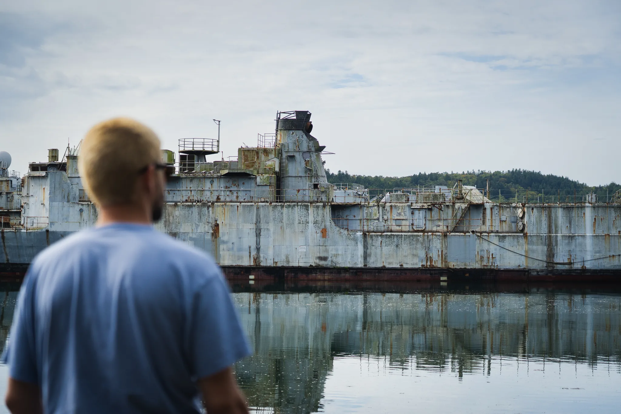 Man in a blue T-shirt seen from behind, looking at decommissioned warships reflected in the still river