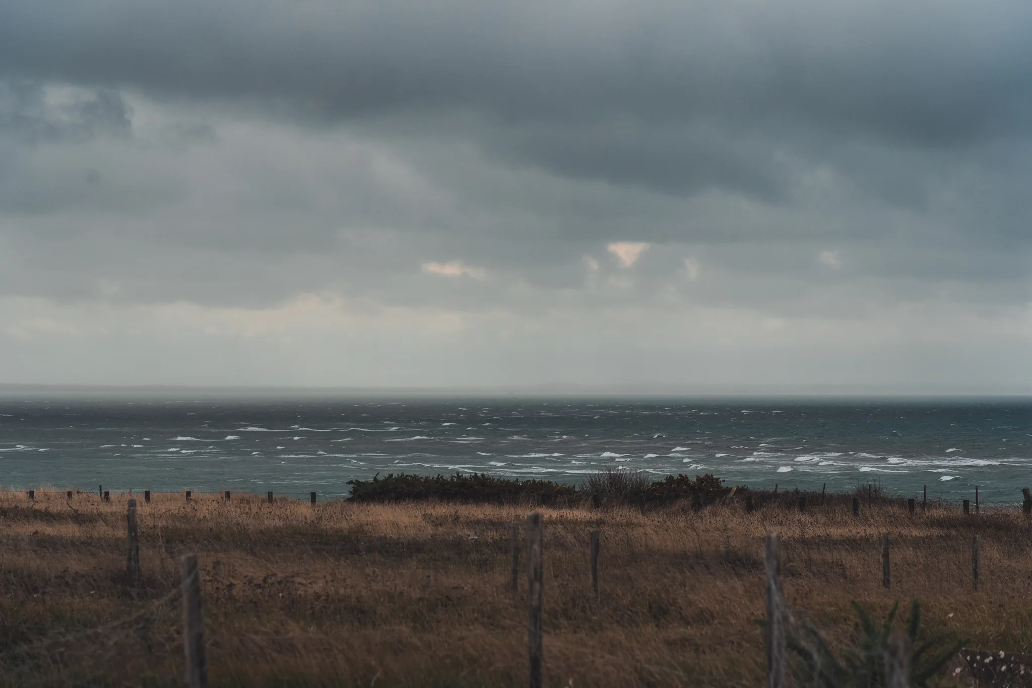 Rough sea beyond a wire fence and brown grass