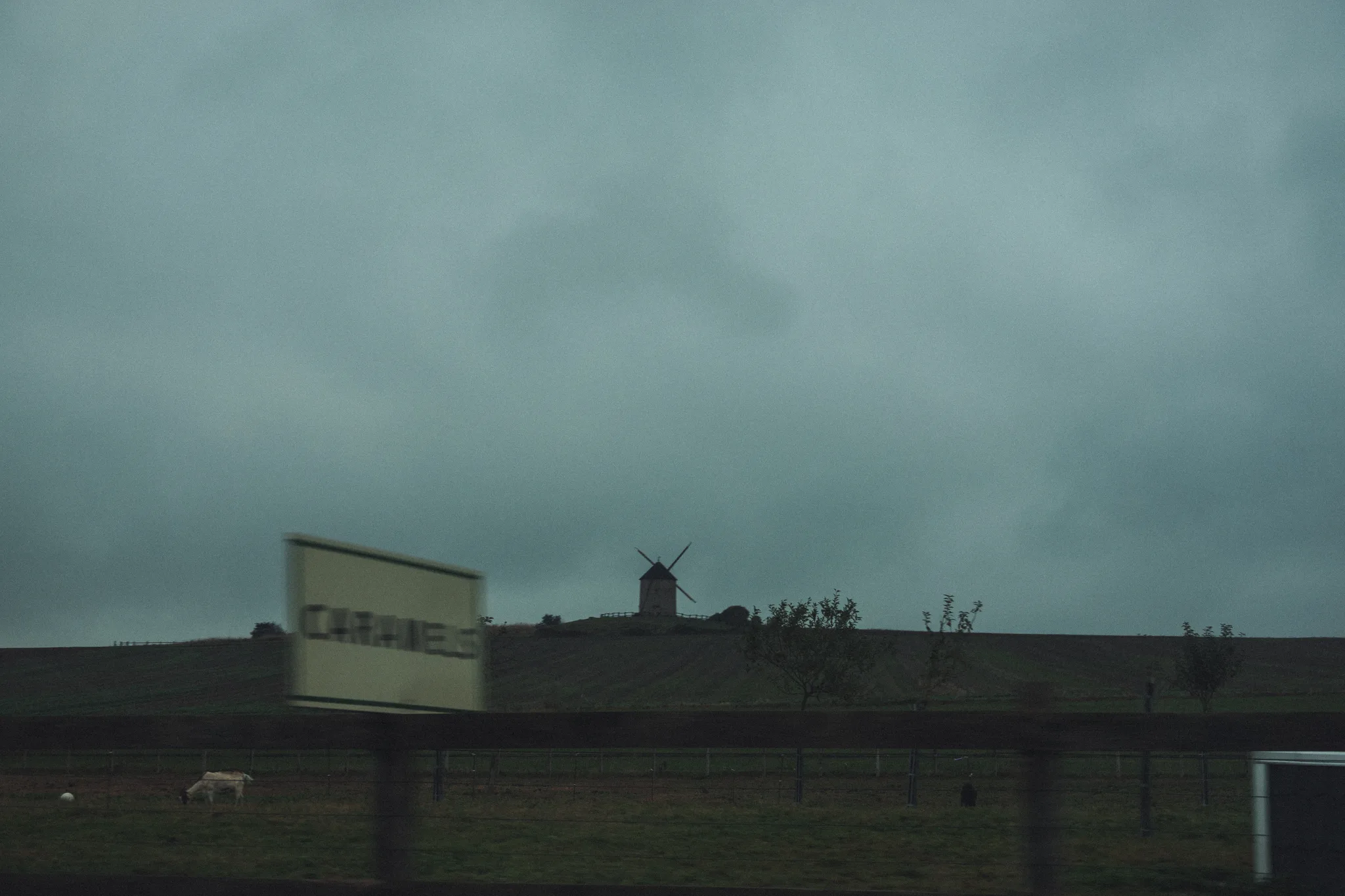 Windmill on a hill and a blurred town sign seen from a moving car under a green-grey sky