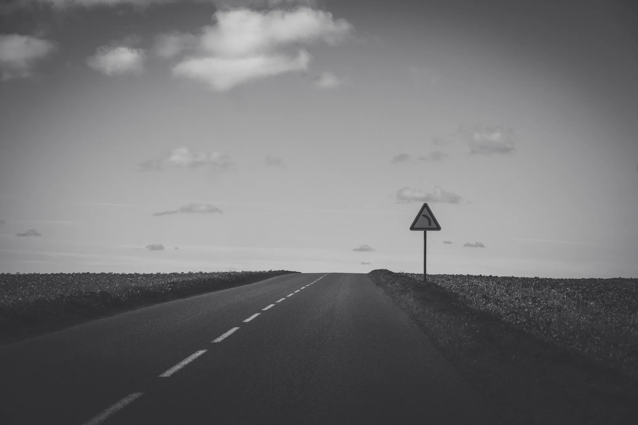 Straight road through fields toward a lone warning sign, black and white